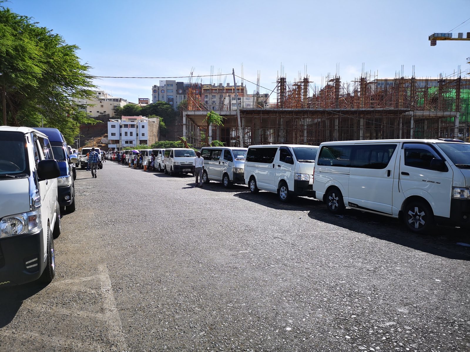 Hilera de furgonetas del transporte colectivo estacionadas en una terminal de Praia, con edificios en construcci&oacute;n al fondo